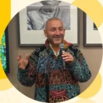 Smiling man in a colorful patterned shirt speaks into a microphone in front of framed photos in an indoor setting.