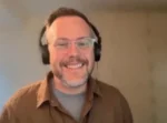 Smiling man with glasses wearing over-ear headphones on a video call, brown shirt over a gray tee.