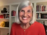 Smiling older woman with short gray hair wearing a red top, indoors with bookshelves in the background.
