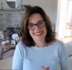 Smiling woman with shoulder-length hair and glasses on a video call, seated in a bright living room with a stone fireplace.