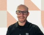Portrait of a smiling man with glasses, wearing a black polo shirt with a small logo, in front of an orange-and-white geometric wall backdrop.