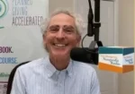 Smiling older man in a light striped shirt speaks into a microphone in a radio studio, with a banner about planned giving in the background.
