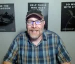 Smiling man with glasses and a cap on a video call, wearing a blue plaid shirt; three framed posters on the wall behind him.