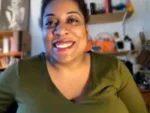 Smiling woman in a green top sits in a home office with shelves behind her.