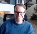 Smiling man with short hair and glasses wearing a blue knit sweater, seated in a bright home office.