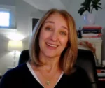 Smiling woman with shoulder-length blonde hair sits in a home office, bookshelves and a plant in the background.