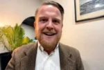 Man in a brown blazer and white shirt smiling during a video call, with a plant and framed photo in the background.
