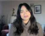 Woman with long dark hair in a brown turtleneck sweater smiles at the camera in a cozy living room with framed art on the wall behind her.