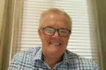 Older man with gray hair and glasses smiling at the camera, wearing a blue checkered shirt in a bright room with blinds on the window.