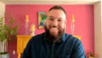 Smiling man with glasses and a full beard sits in a pink-walled room, wearing a blue button-up shirt.