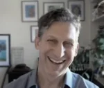 Smiling man with short gray hair wearing a light blue shirt, in a home office with framed photos and plants in the background.