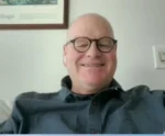 Smiling bald man with round glasses on a video call, wearing a dark shirt, wireless earbuds visible, in a room with a framed picture.