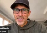 Smiling man wearing a dark cap and glasses during a video call, indoors; name label partially visible in the corner.