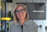 Smiling woman with blonde hair and large glasses speaking on a Bloomberg video backdrop.
