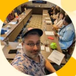 Selfie of a man wearing glasses and a cap at a conference room table, with several people seated around and papers and drinks on the table behind him.