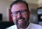 Smiling man with round glasses and a beard, wearing a white shirt in an office setting.