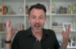 Man in a black shirt speaks excitedly into a microphone, gesturing with both hands in front of a bookshelf backdrop.