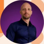 Professional headshot of a bald man in a navy button-down shirt against a purple gradient backdrop.
