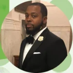 Portrait of a man in a black tuxedo with a bow tie and white flower boutonniere, standing indoors.