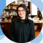Professional woman with a black blazer and red glasses smiling in a warmly lit bookstore or café background (head-and-shoulders).