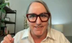Woman with red chunky glasses smiling at camera during a video call in a home office setting.