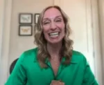 Smiling woman with wavy hair wearing a green blouse, facing the camera in a home setting with framed wall art behind her.
