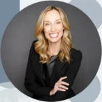 Professional headshot of a smiling blonde woman in a black blazer against a gray backdrop.