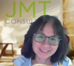 Smiling woman with short dark hair and glasses, in an office setting with a JMT CONSULT sign in the background.