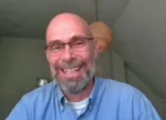 Smiling bald man with glasses and gray beard in a blue shirt on a video call.