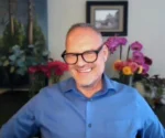 Smiling man in a blue shirt and black-framed glasses, indoors with colorful flowers in the background.