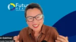 Smiling woman with short gray hair and glasses speaks at a virtual event, wearing a brown blouse with a microphone, Futuro Health branding in the blue background behind her.