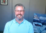 Smiling man with gray hair and beard wearing glasses and a light blue polo, standing indoors with a gray wall and a brochure display in the background.