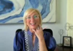 Smiling blonde woman in a blue blouse and glasses sits at a desk, speaking on a video call with abstract art in the background.
