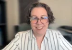 Smiling woman with short, curly hair and glasses wearing a white striped shirt during a video call in a home/office setting