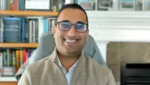 Smiling man wearing glasses, seated in a home office with a bookshelf in the background.