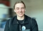Person smiling, wearing a dark zip-up jacket with a circular badge, indoors.