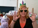 Woman with blonde hair wearing a unicorn headband and heart-shaped sunglasses, hands raised in a playful pose in a toy-filled room.