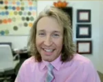 Smiling man with shoulder-length hair in a pink shirt and striped tie, in an office with color swatches on the wall.