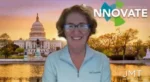Smiling woman presenter in a light blue polo stands in front of a sunset backdrop with the Capitol and Innovate branding.