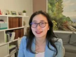 Smiling woman with round glasses on a video call, wearing a light blue blazer in a home office with shelves behind her.
