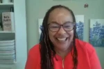Smiling woman with glasses and braided hair wearing a red top, indoors.