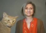 Smiling woman in an orange blouse and brown cardigan standing beside an orange tabby cat against a gray background.