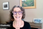 Smiling woman with curly gray hair and pink glasses on a video call; desk behind her with framed art and a plate reading 'NPOGURU'.