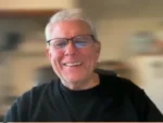 Smiling older man with gray hair and glasses, wearing a black shirt, looking at the camera indoors with a blurred background.
