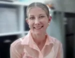 Smiling woman with light hair in a pink blouse and pearl earrings, in an office setting.