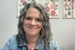 Smiling woman with shoulder-length curly gray hair wearing a denim jacket, indoors with floral artwork on the wall behind her.