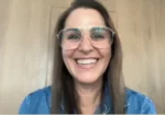 Smiling woman with long hair and large glasses, wearing a blue denim shirt, on a video call.