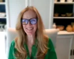 Woman with long blonde hair and blue-framed glasses smiling at the camera in a green blouse, home setting in background.