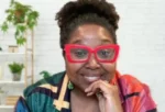 Woman smiling at the camera with bright red rectangular glasses and a colorful striped shirt; plant and white wall background behind her.