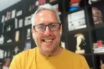 Smiling man with gray hair and glasses wearing a yellow shirt, in a room with dark shelving and assorted items in the background.